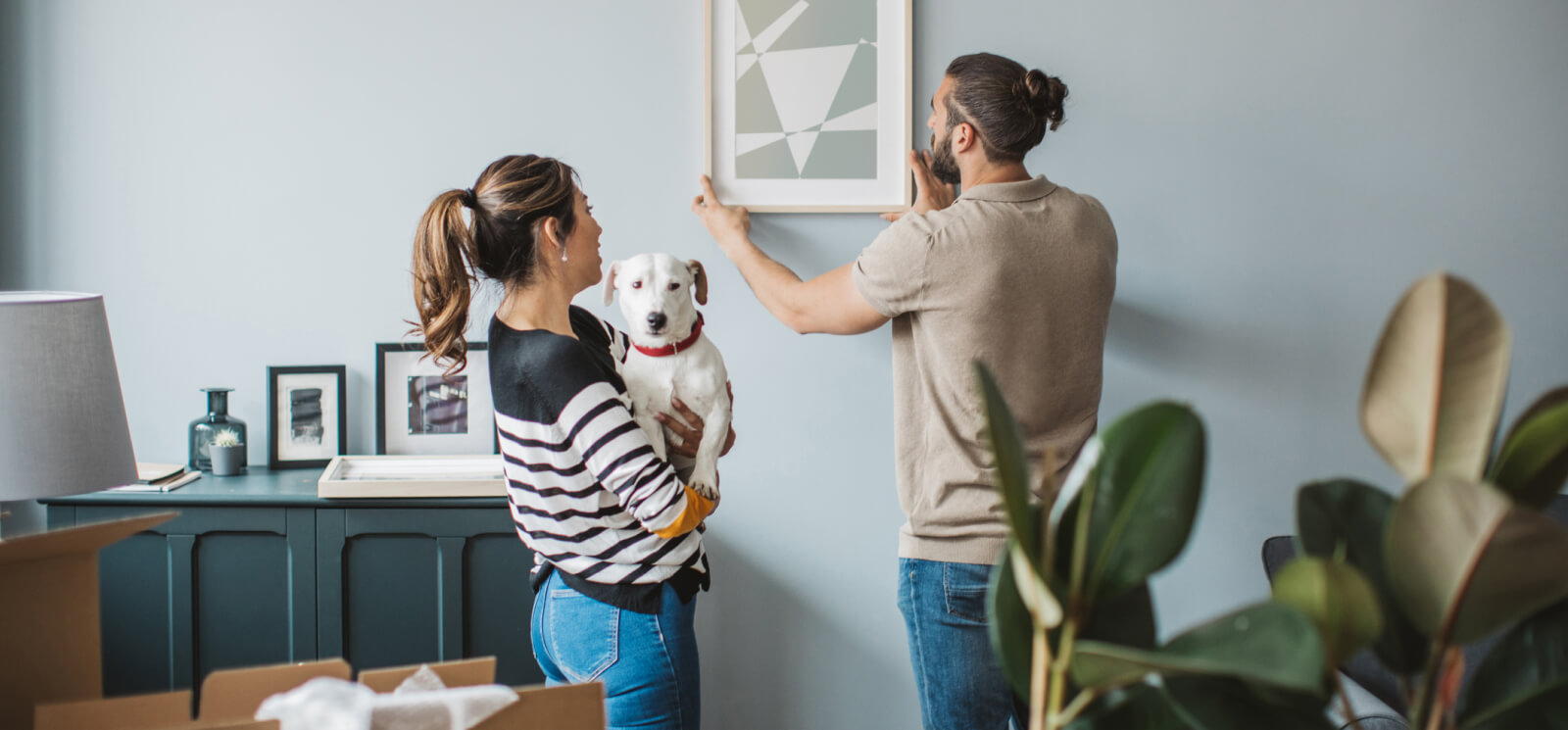 A young couple and a small dog hanging a framed piece of art in a living room