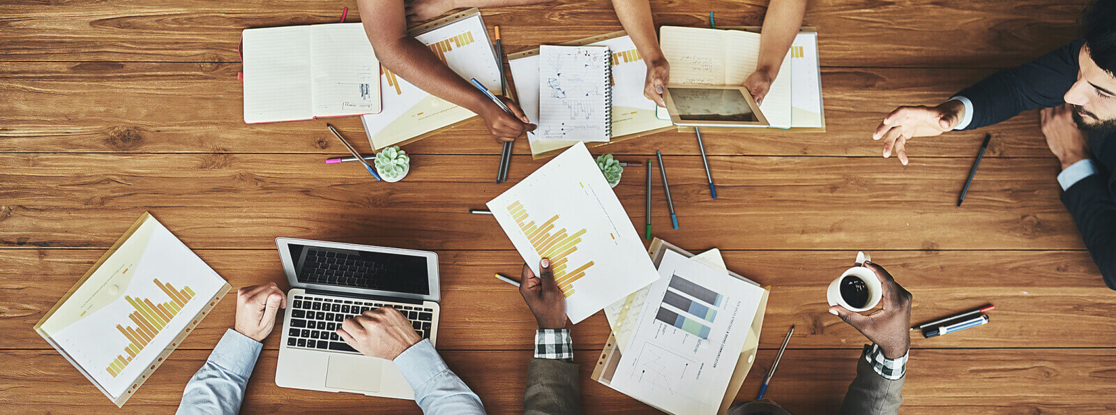 Top-down view of group of business people around a long desk with documents and laptops